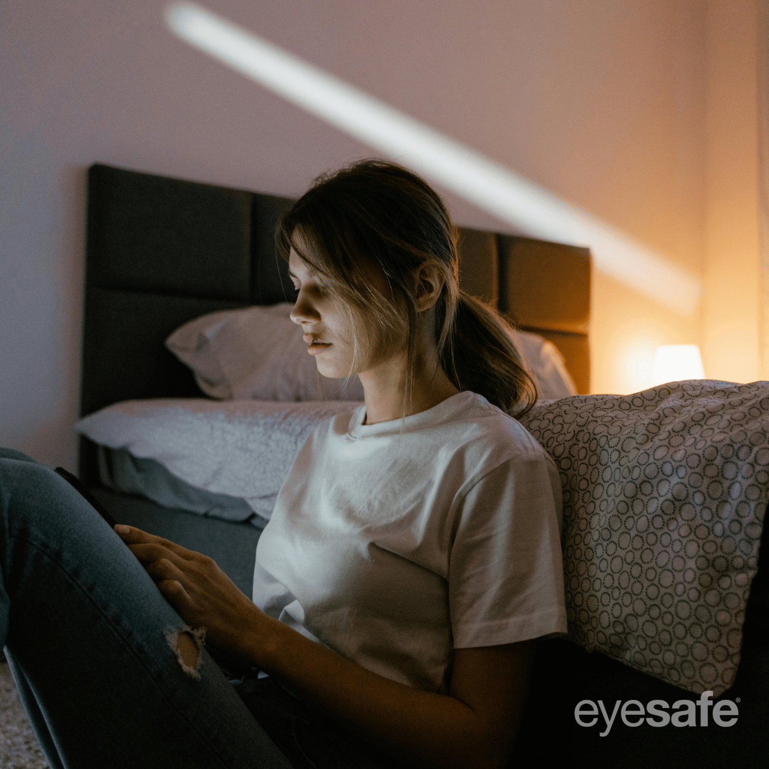 oung woman sitting on the floor at night using a smartphone, bedroom lighting in the background, Eyesafe logo visible.