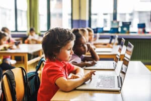 Group of elementary school students sitting at desks in a classroom using laptops for learning. A young boy in a red shirt is focused on the screen, with other students working in the background.